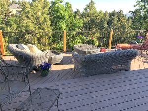 Two-level wooden deck surrounded by trees. Deck is enclosed by a metal railing. Lower level of deck has rattan seating and table with chaise lounge chair and potted flowers.