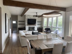 Remodeled living dining area with a window wall, floor to ceiling stone fireplace surround for fireplace, ledge and large flat screen TV. On both sides of wall are built in floating shelves and cabinets. High ceilings include wooden beams, a large dining room modern circular chandelier.