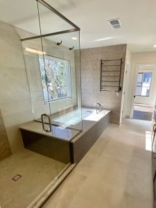 Large modern bathroom remodeling that includes two-sided glass shower walls, a separate granite-surround bathtub with large window showing view of tree. Wall at end of tub features popular herringbone tile pattern with metal towel rack. Granite countertop with two sinks sits across from shower and tub.